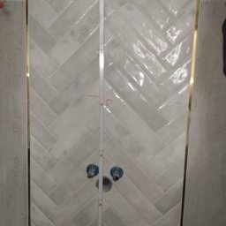 EDE GROUP SPÓŁKA Z OGRANICZONĄ ODPOWIEDZIALNOŚCIĄ - Ułożone białe płytki w jodełkę z ozdobnymi listwami w łazience w Słubicach. Widoczne przyłącza wodne i przymiar. Nietypowy wzór płytek.