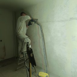 EDE GROUP SPÓŁKA Z OGRANICZONĄ ODPOWIEDZIALNOŚCIĄ - Pracownik na drabinie wycina bruzdy w ścianie pod instalację elektryczną za pomocą frezarki z odkurzaczem. Widoczne przewody i zarys instalacji.