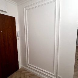 EDE GROUP SPÓŁKA Z OGRANICZONĄ ODPOWIEDZIALNOŚCIĄ - Biała ściana z dekoracyjną ramą i listwami przypodłogowymi, brązowe drzwi po lewej stronie, parkiet ułożony w jodełkę. Nowoczesne wykończenie wnętrza.