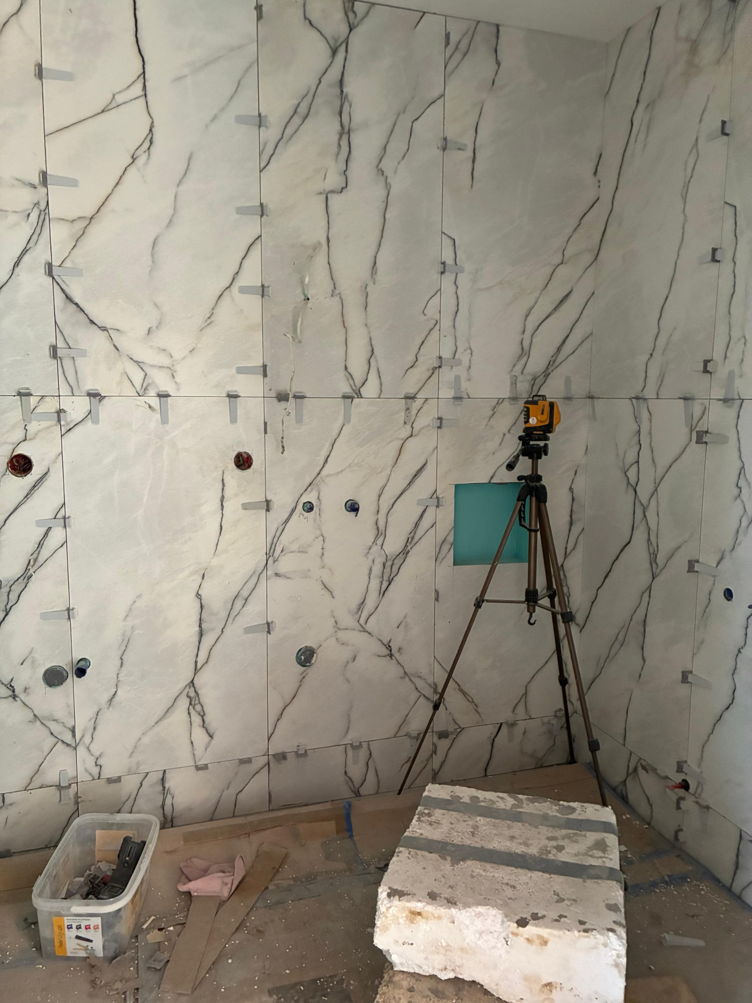 Układanie płytek imitujących marmur w łazience, widoczne poziomice laserowe, krzyżyki dystansowe i narzędzia glazurnicze. Etap pośredni prac.