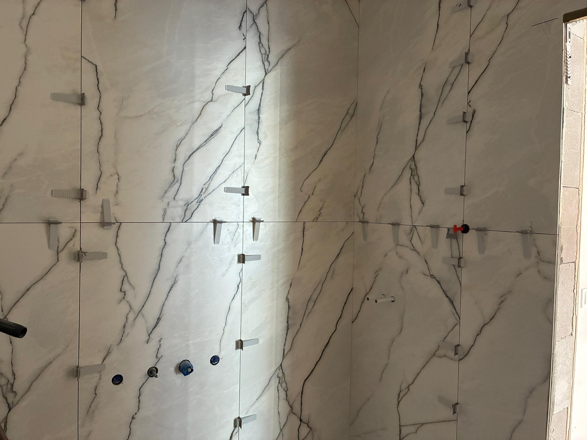 Białe płytki imitujące marmur z czarnymi żyłami, ułożone na ścianie, z widocznymi plastikowymi krzyżykami dystansowymi. Częściowo wykończona ściana.
