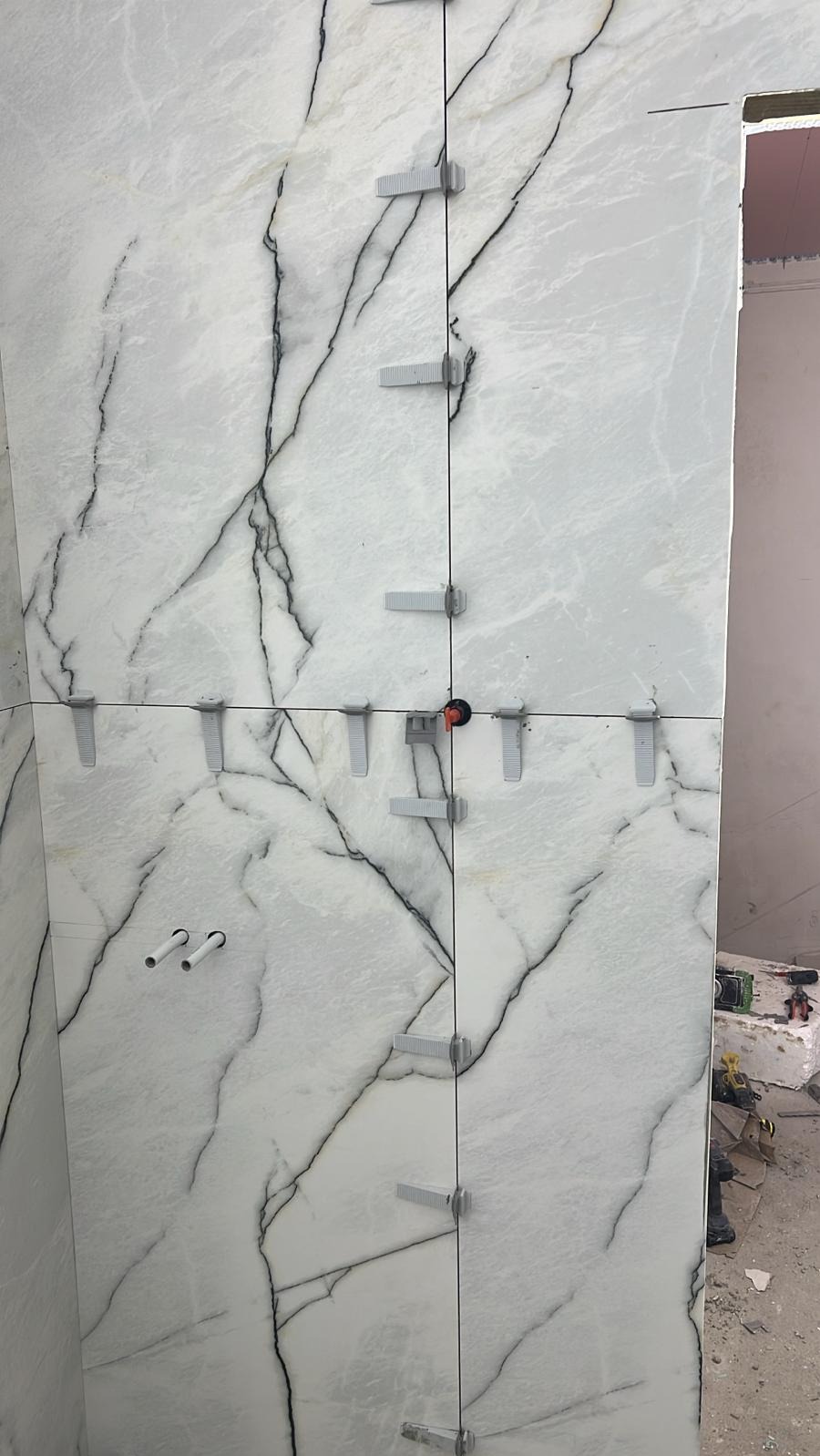 Ściana wyłożona białymi płytkami imitującymi marmur z czarnymi żyłami. Widoczne klipsy poziomujące płytki. Dwa otwory na rury w dolnej części ściany.