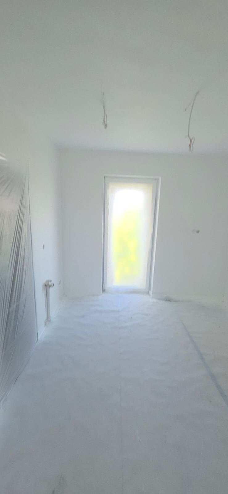 Białe, świeżo pomalowane wnętrze pokoju z zakrytymi folią oknami i podłogą. Widoczne wystające kable z sufitu. Przygotowanie do dalszych prac wykończeniowych.