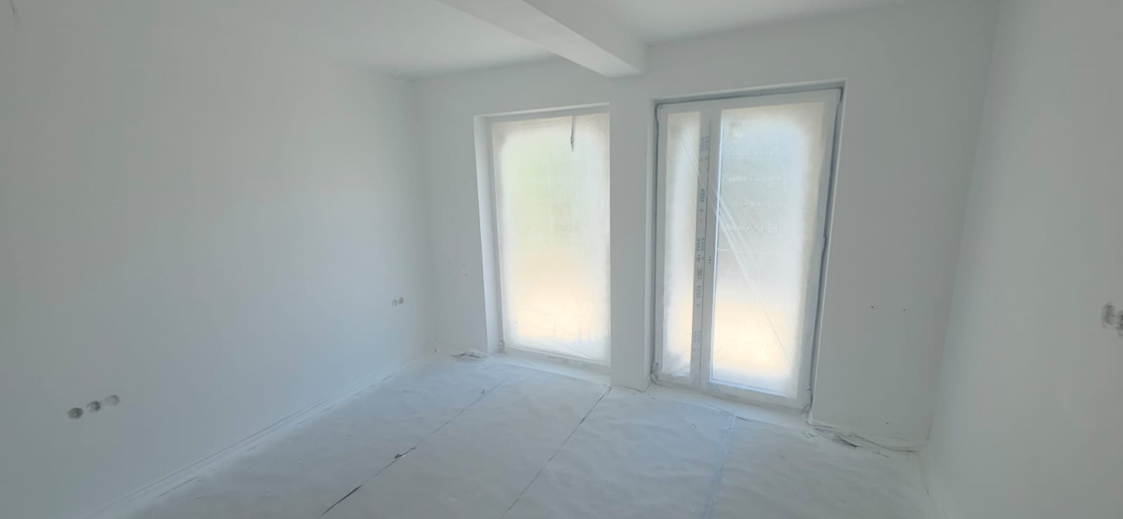 Białe, świeżo pomalowane wnętrze pokoju z dwoma oknami zabezpieczonymi folią malarską i zakrytym podłożem, widoczne gniazdka elektryczne.