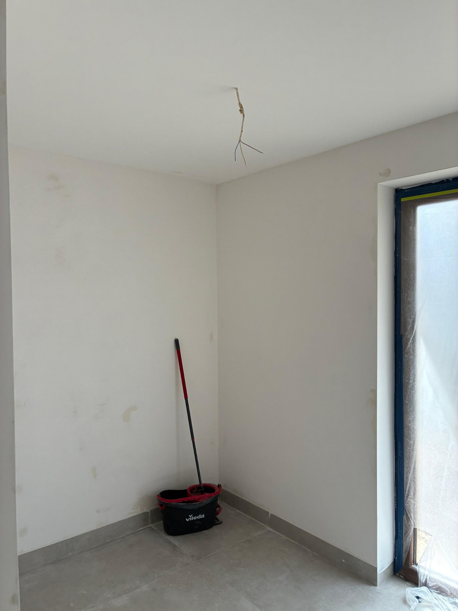Wnętrze w trakcie prac wykończeniowych: białe ściany z plamami, szare płytki podłogowe, wiadro i mop Vileda, odsłonięte przewody elektryczne.