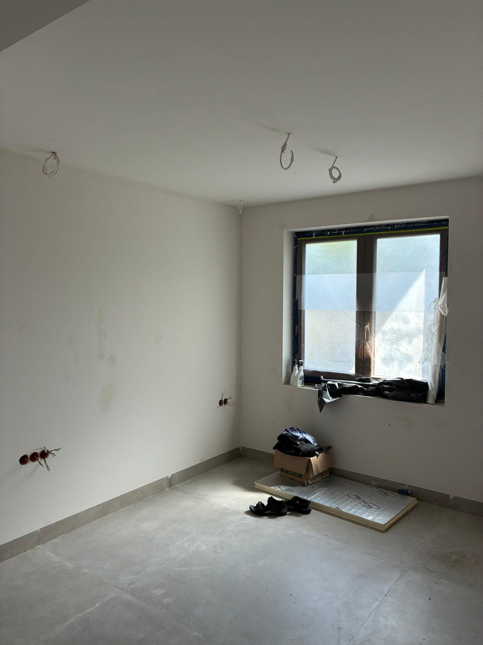 Pokój w trakcie remontu: szare płytki podłogowe, białe ściany z widocznymi puszkami elektrycznymi, okno zabezpieczone folią. Materiały budowlane na podłodze.