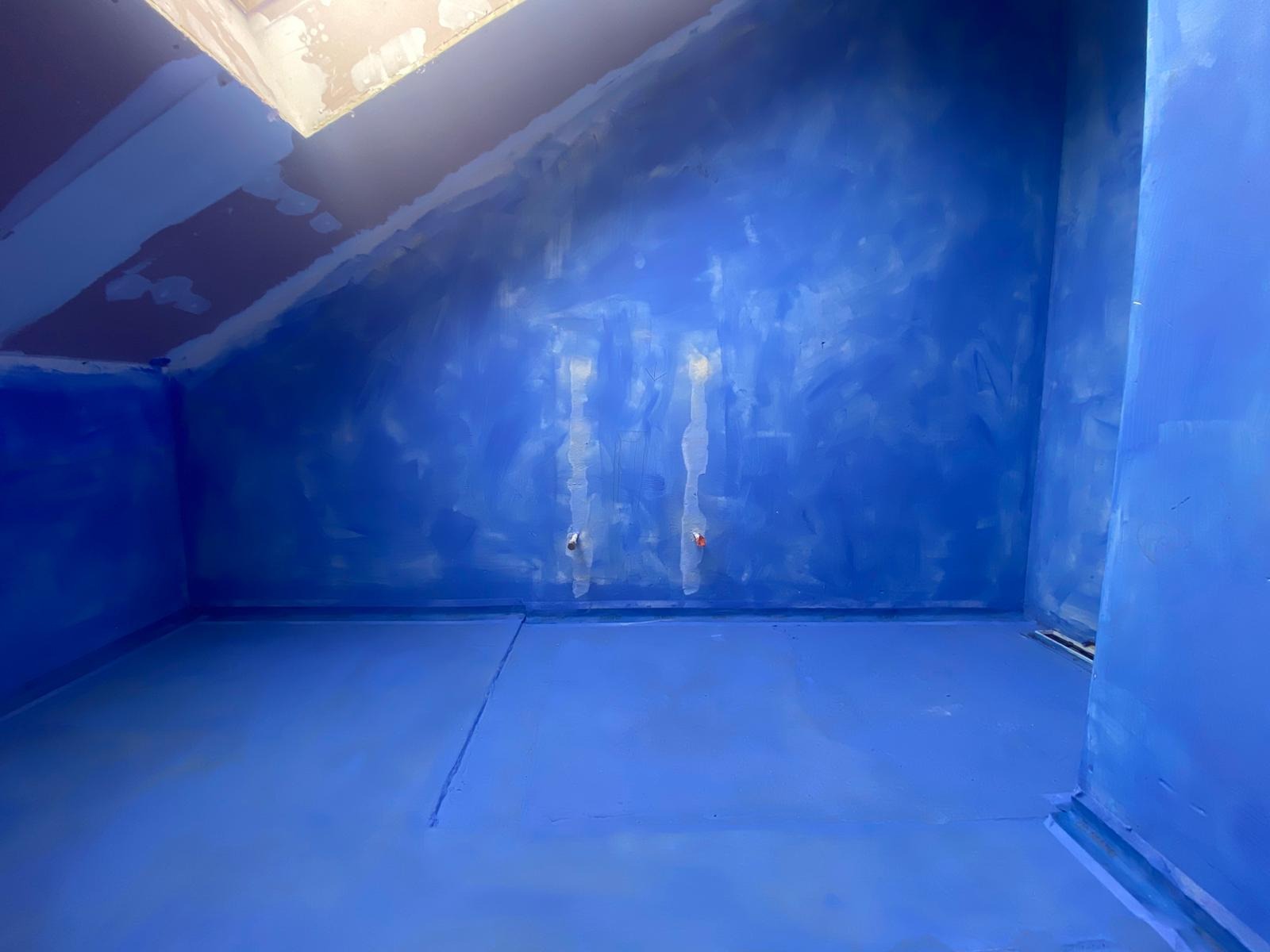 Niebieska hydroizolacja w łazience na poddaszu, widoczne przejścia rur i odpływ liniowy. Przygotowanie do dalszych prac wykończeniowych.