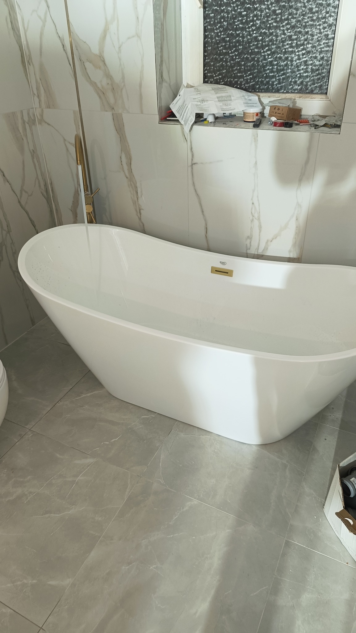 Biała, wolnostojąca wanna wypełniona wodą, z złotą armaturą, na tle marmurowych płytek ściennych i szarych płytek podłogowych. Widoczne narzędzia na parapecie okna.