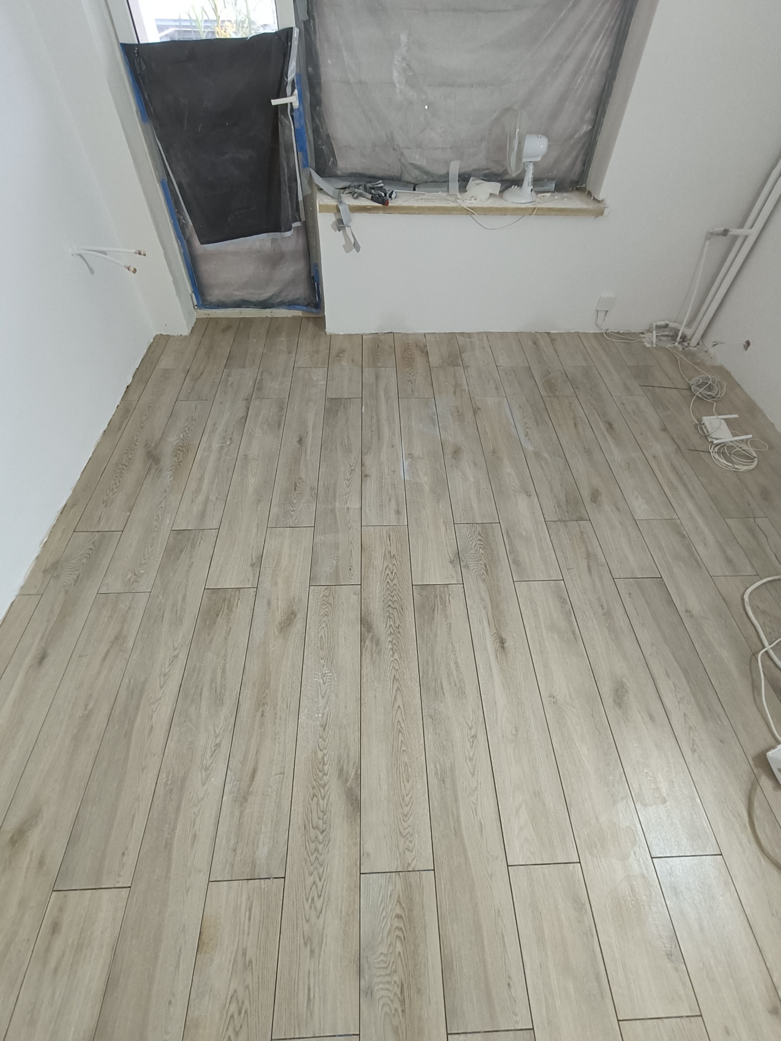 Ułożona podłoga z paneli imitujących drewno w jasnym odcieniu, widok na okno zabezpieczone folią malarską, instalacja elektryczna przy ścianie.