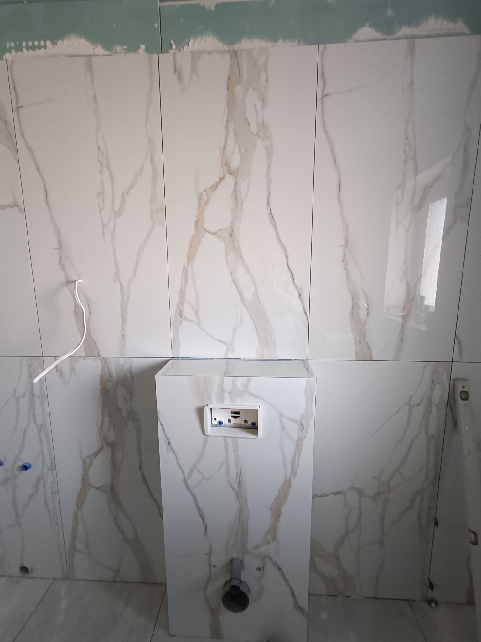 Łazienka w trakcie wykańczania: białe płytki imitujące marmur na ścianach, zabudowa stelaża WC, widoczne elementy instalacji. Poziomica przy ścianie.