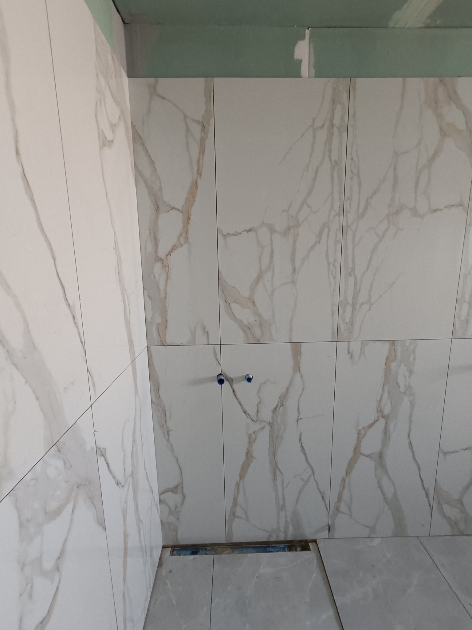 Ściana i podłoga w trakcie wykańczania płytkami imitującymi marmur. Widoczne niebieskie elementy instalacji i fragment surowego sufitu. Jasne, neutralne barwy.