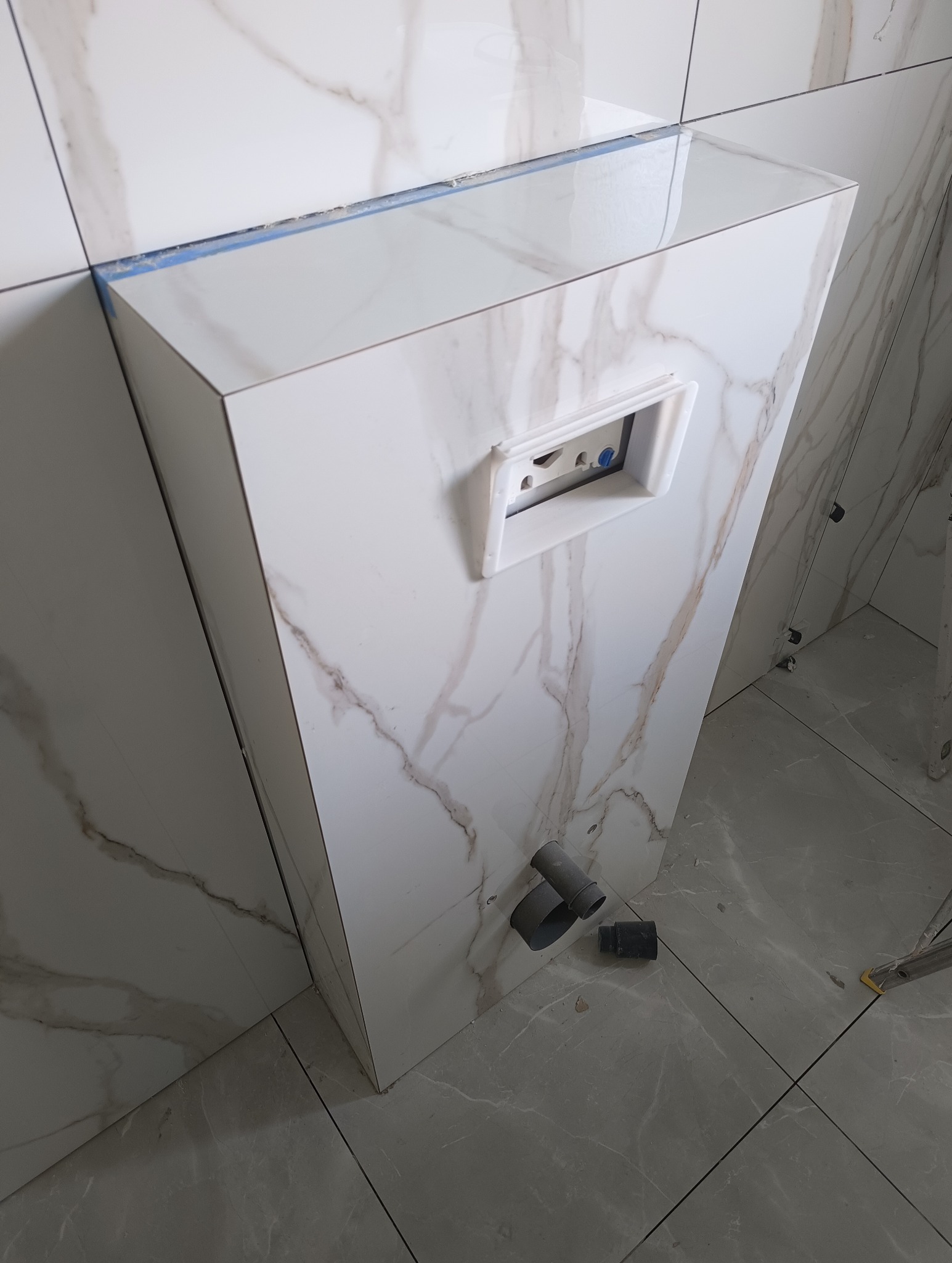 Obudowa stelaża WC z płytek imitujących marmur, z widocznym otworem na przycisk spłukujący i rurami odpływowymi na podłodze. Ukończona zabudowa w trakcie prac wykończeniowych.