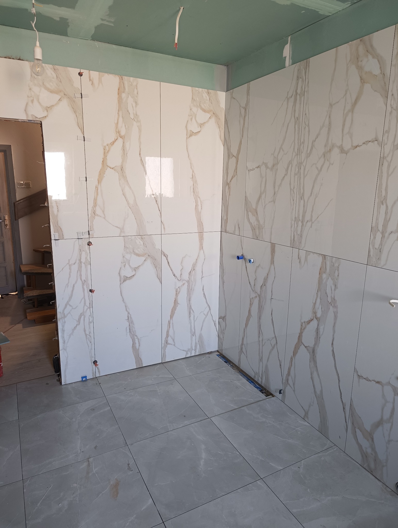 Łazienka w trakcie wykańczania: marmurowe płytki na ścianach z widocznymi krzyżykami dystansowymi, szare płytki podłogowe. Widoczne niedokończone elementy instalacji.