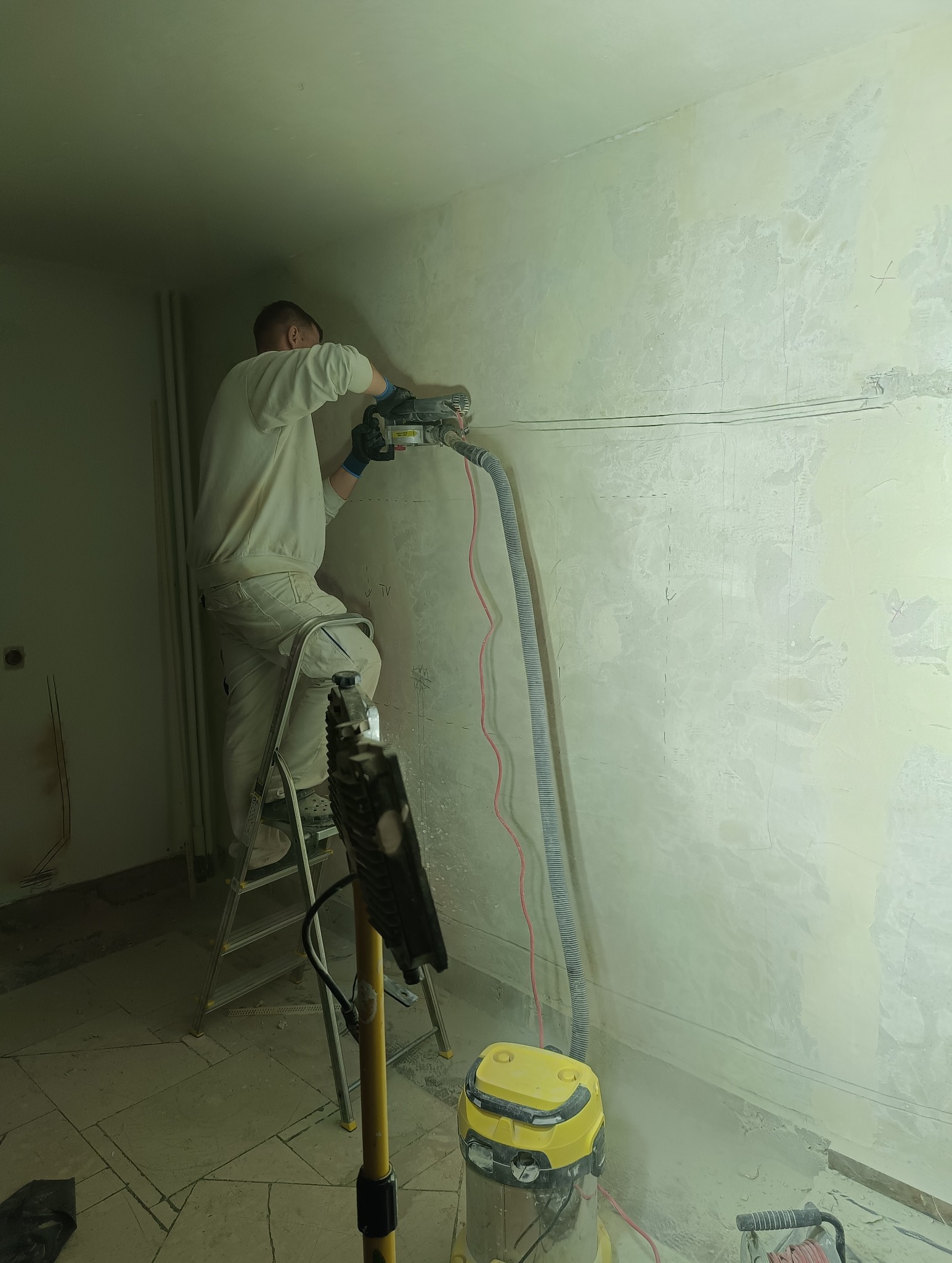 Pracownik na drabinie wycina bruzdy w ścianie pod instalację elektryczną za pomocą frezarki z odkurzaczem. Widoczne przewody i zarys instalacji.