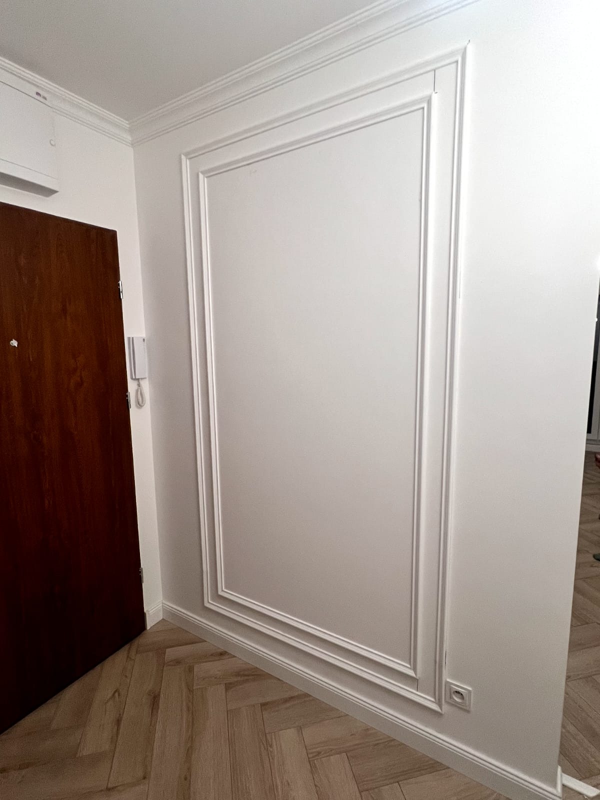 Biała ściana z dekoracyjną ramą i listwami przypodłogowymi, brązowe drzwi po lewej stronie, parkiet ułożony w jodełkę. Nowoczesne wykończenie wnętrza.