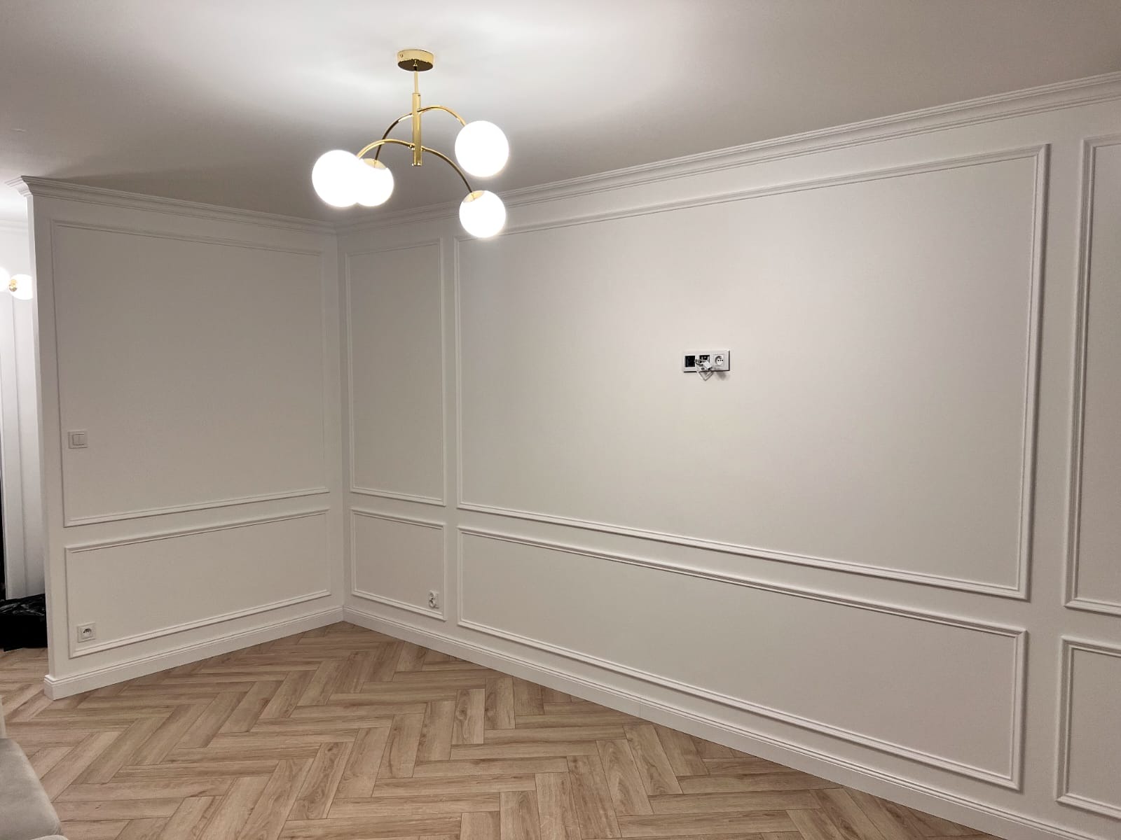 Eleganckie wykończenie wnętrza: białe ściany z dekoracyjnymi listwami, parkiet w jodełkę i nowoczesna, złota lampa sufitowa z białymi kulami.