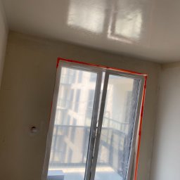 Spray-Tech - Pokój w trakcie prac wykończeniowych, okno zabezpieczone folią i taśmą, ściany przygotowane pod malowanie lub tapetowanie, widoczna gładka powierzchnia sufitu.