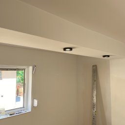 Spray-Tech - Wnętrze w trakcie prac wykończeniowych we Wrocławiu: ściana po szpachlowaniu i malowaniu, widoczne dwa okrągłe punkty świetlne w suficie oraz poziomica oparta o ścianę.