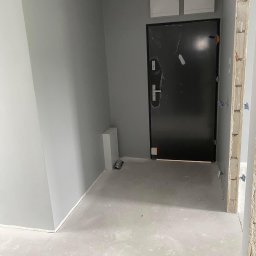 Spray-Tech - Wrocław: Świeżo pomalowane na szaro ściany w przedpokoju mieszkania, z widoczną czarną, oklejoną folią ochronną, metalową futryną drzwi wejściowych i betonową wylewką na podłodze.