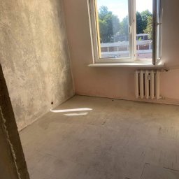 Spray-Tech - Pokój w trakcie przygotowań do prac wykończeniowych: ściany po wstępnym szpachlowaniu, zabezpieczona podłoga, widoczny grzejnik i otwarte okno z widokiem na drzewa.