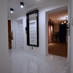 Nowoczesny przedpokój z lustrem w czarnej ramie, białą marmurową podłogą i widokiem na salon z ceglaną ścianą i parkietem w jodełkę.