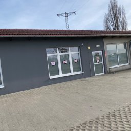 Spray-Tech - Szary budynek z białymi oknami i drzwiami, z plakatami w oknach, na tle błękitnego nieba i słupa wysokiego napięcia.