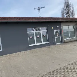 Szary budynek z białymi oknami i drzwiami, z plakatami w oknach, na tle błękitnego nieba i słupa wysokiego napięcia.