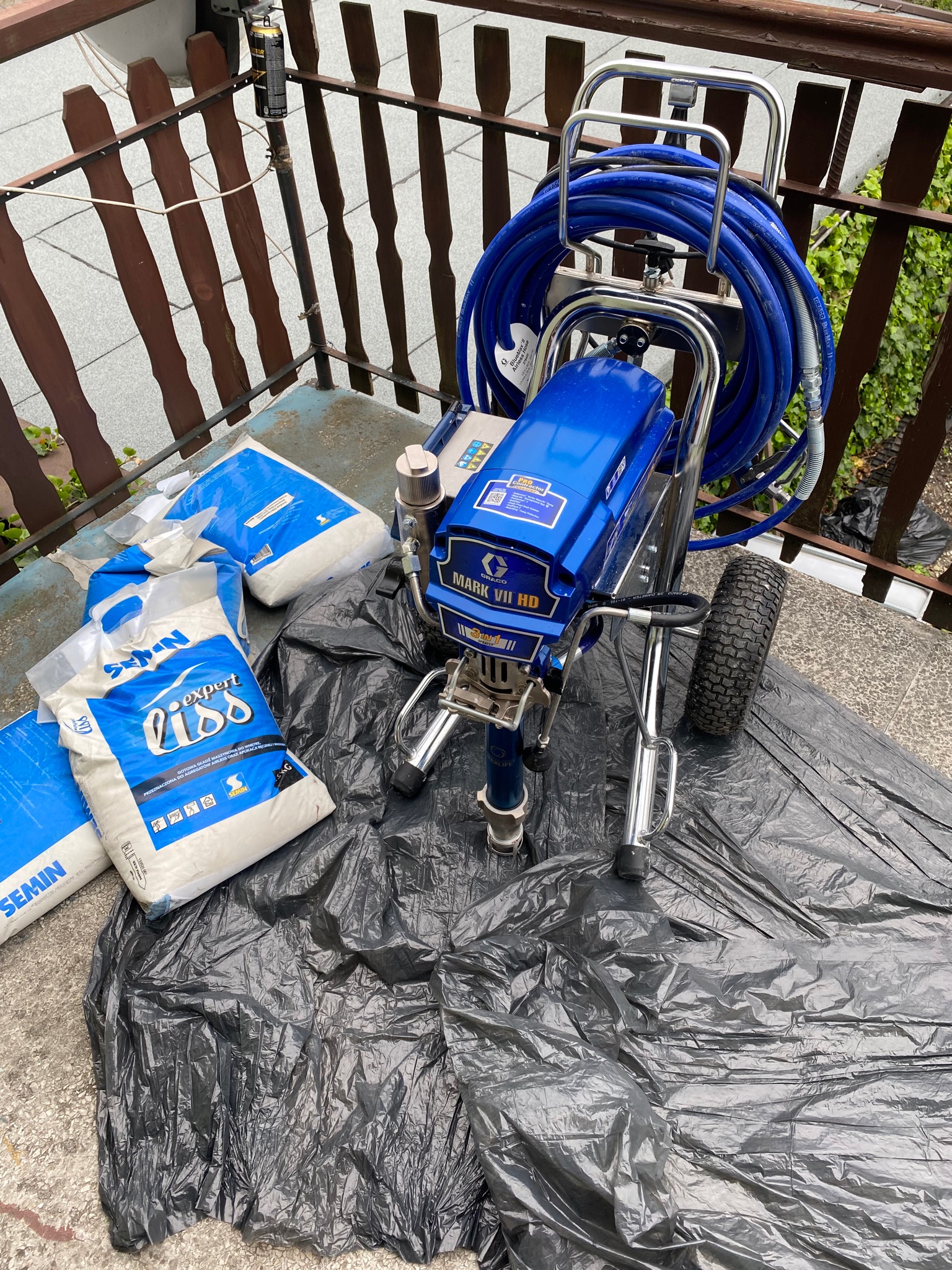 Agregat malarski Graco Mark VII HD z niebieskim wężem i workami gotowej masy szpachlowej Semnin Expert Liss na czarnej folii ochronnej na balkonie.