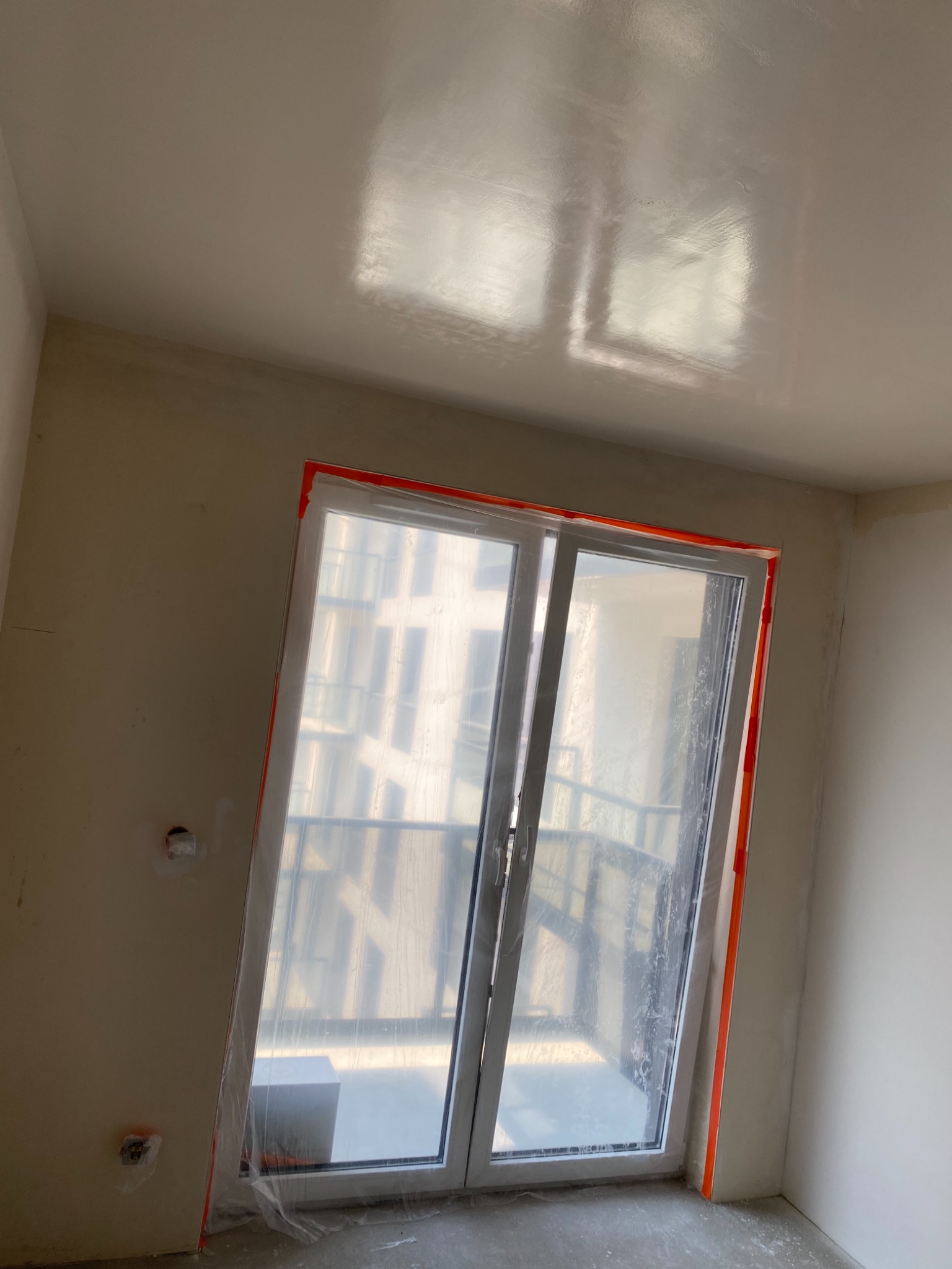 Pokój w trakcie prac wykończeniowych, okno zabezpieczone folią i taśmą, ściany przygotowane pod malowanie lub tapetowanie, widoczna gładka powierzchnia sufitu.