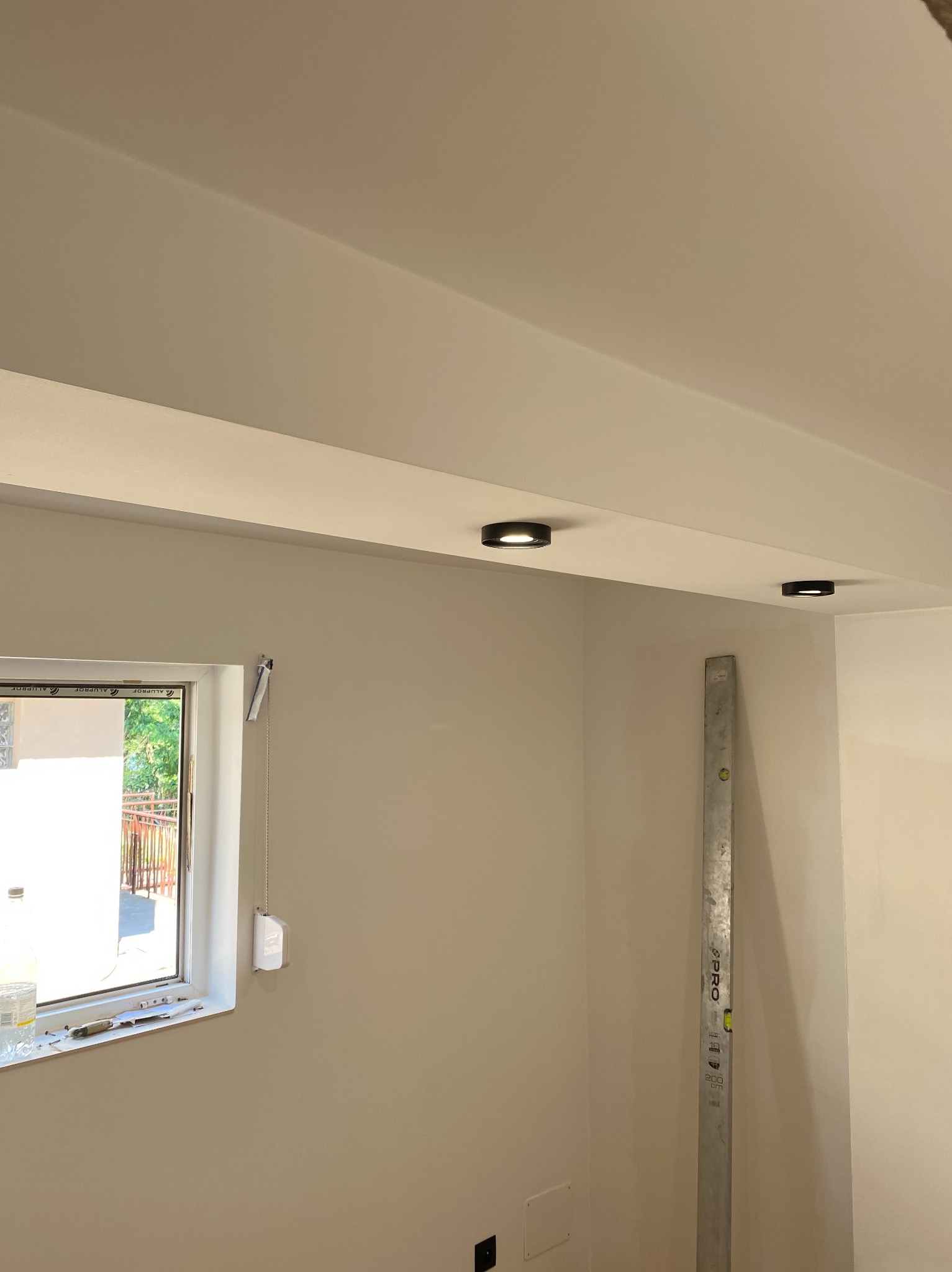 Wnętrze w trakcie prac wykończeniowych we Wrocławiu: ściana po szpachlowaniu i malowaniu, widoczne dwa okrągłe punkty świetlne w suficie oraz poziomica oparta o ścianę.