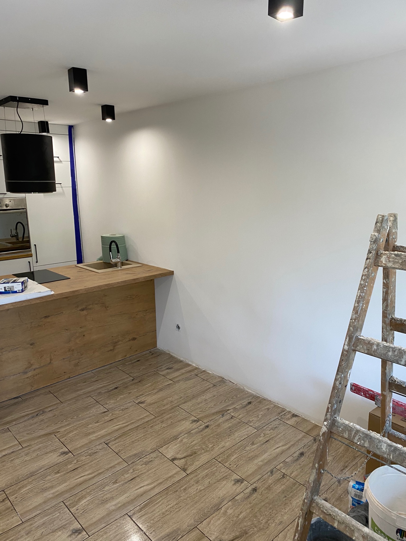 Świeżo pomalowana na biało ściana w kuchni z drewnianą zabudową, widoczna drabina malarska z resztkami farby i wiadro z farbą, podłoga wyłożona płytkami imitującymi drewno.