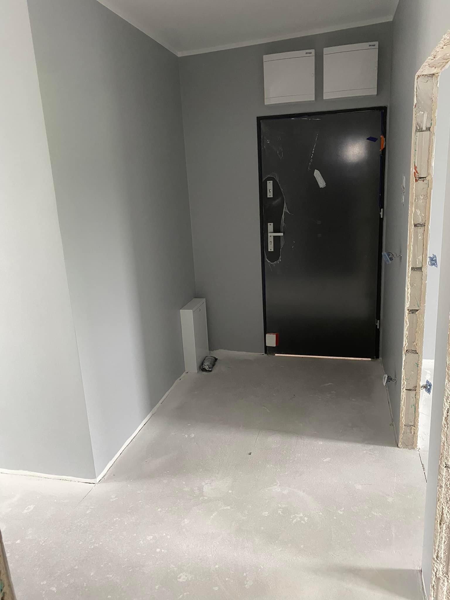 Wrocław: Świeżo pomalowane na szaro ściany w przedpokoju mieszkania, z widoczną czarną, oklejoną folią ochronną, metalową futryną drzwi wejściowych i betonową wylewką na podłodze.