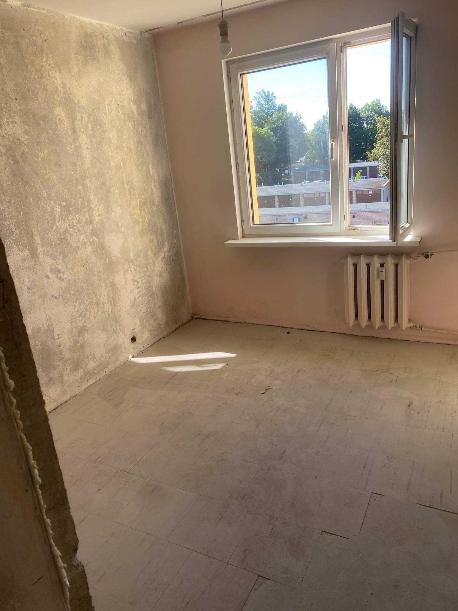 Pokój w trakcie przygotowań do prac wykończeniowych: ściany po wstępnym szpachlowaniu, zabezpieczona podłoga, widoczny grzejnik i otwarte okno z widokiem na drzewa.