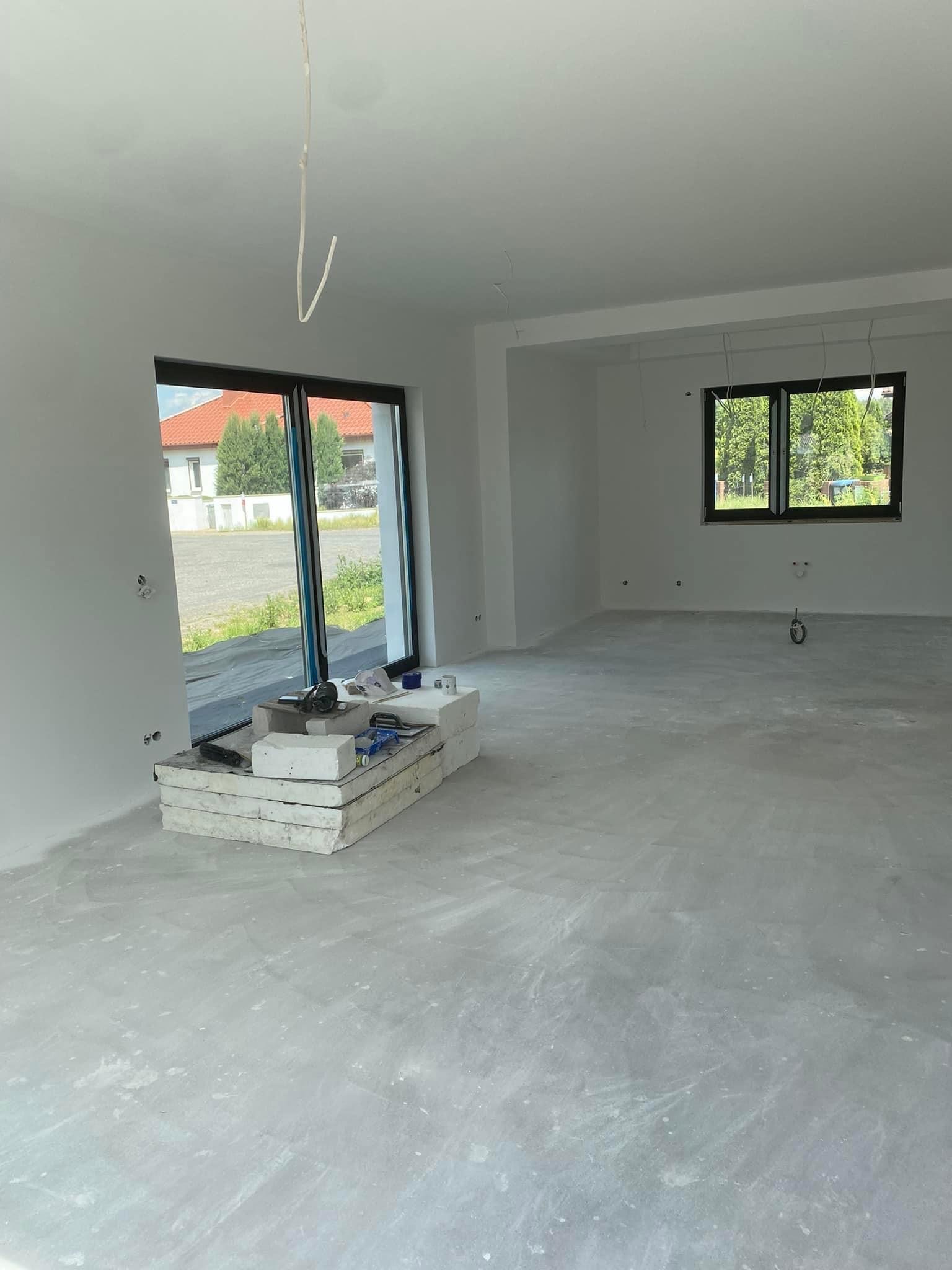 Wnętrze w stanie deweloperskim, z oknami, świeżo wylaną wylewką podłogową i przygotowanymi narzędziami przy oknie tarasowym.