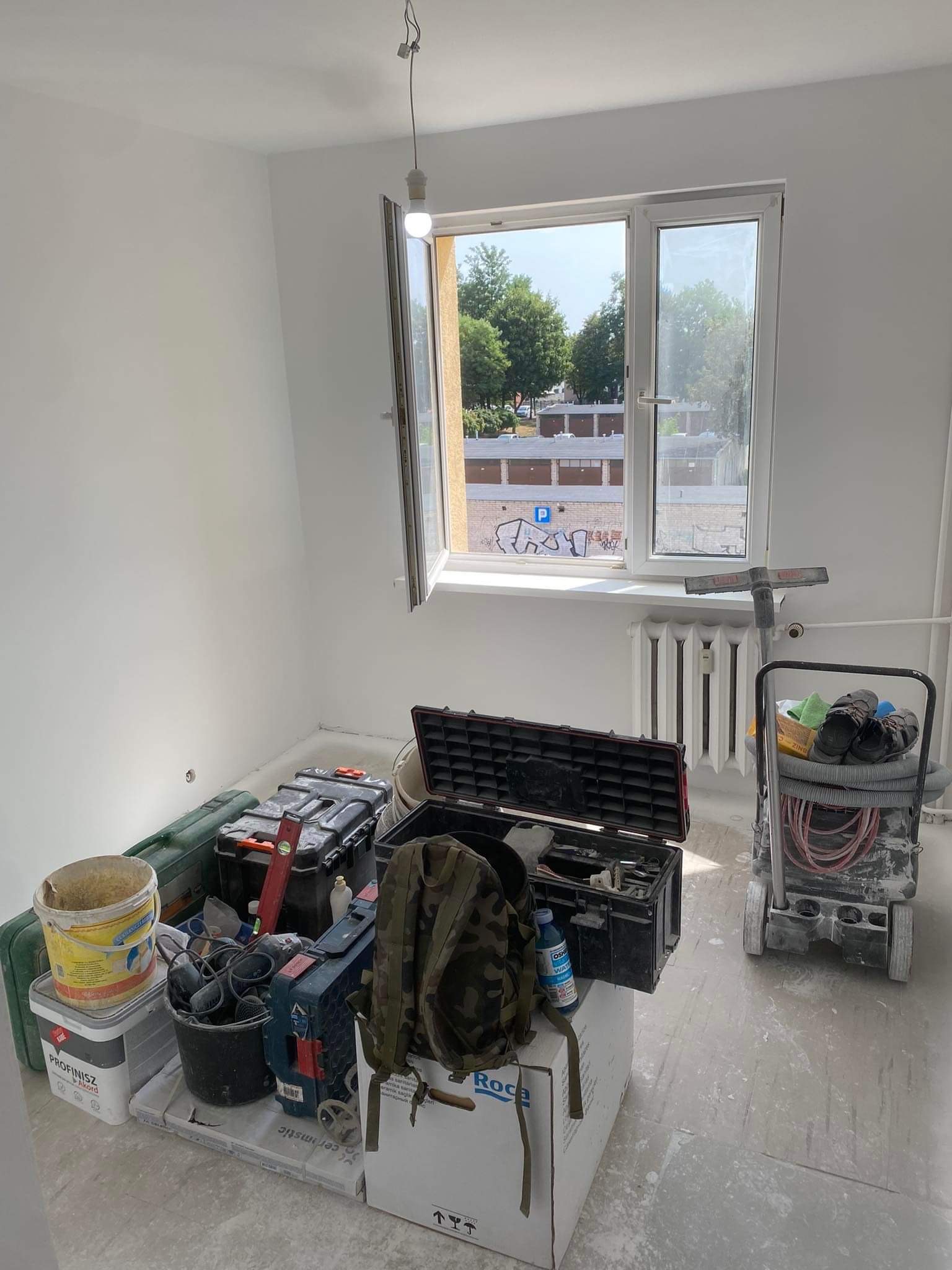 Pokój w trakcie remontu: ściany świeżo pomalowane na biało, okno otwarte z widokiem na budynki, narzędzia i materiały budowlane porozrzucane na podłodze, widoczny agregat malarski i skrzynki...