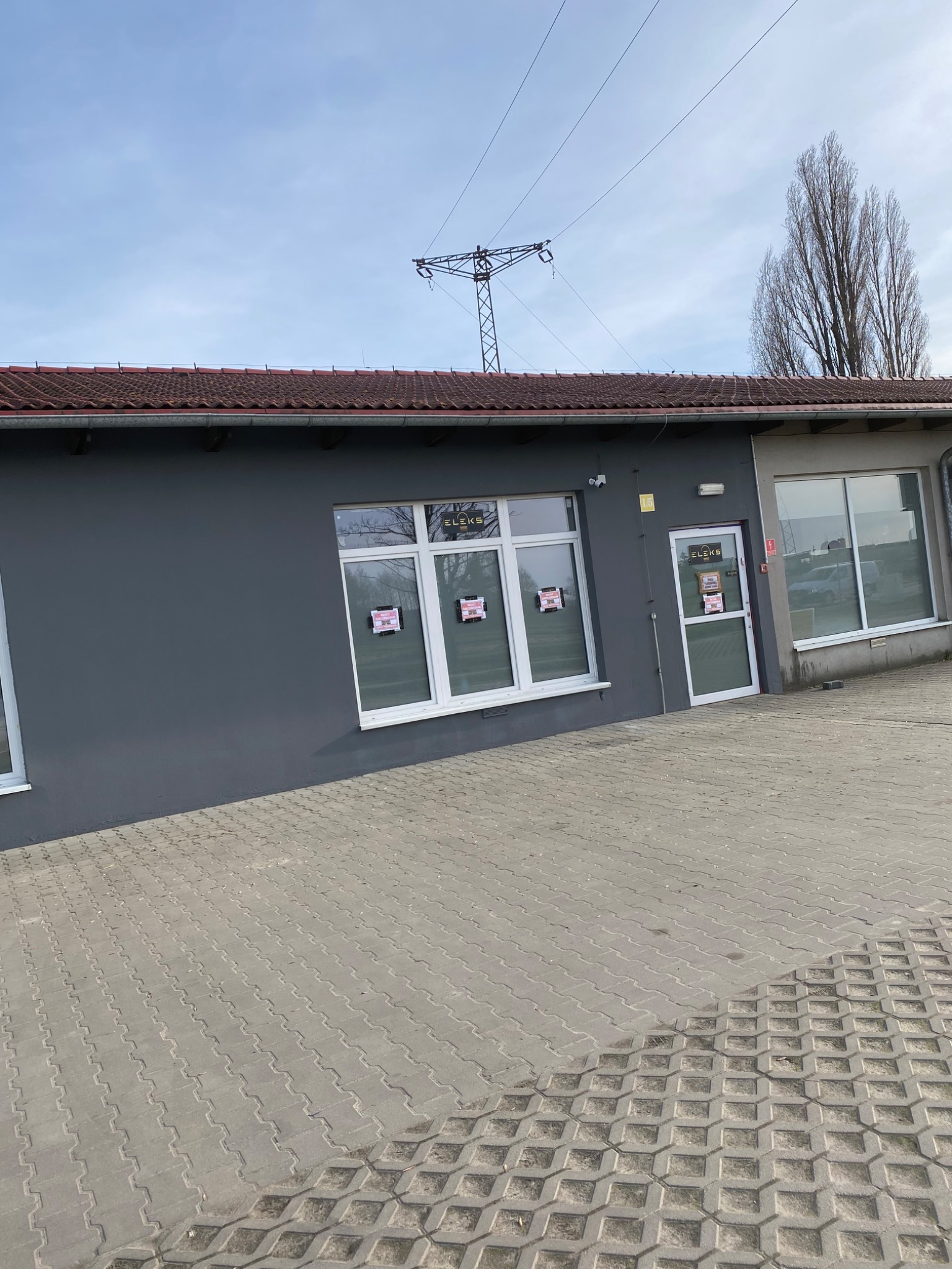 Szary budynek z białymi oknami i drzwiami, z plakatami w oknach, na tle błękitnego nieba i słupa wysokiego napięcia.