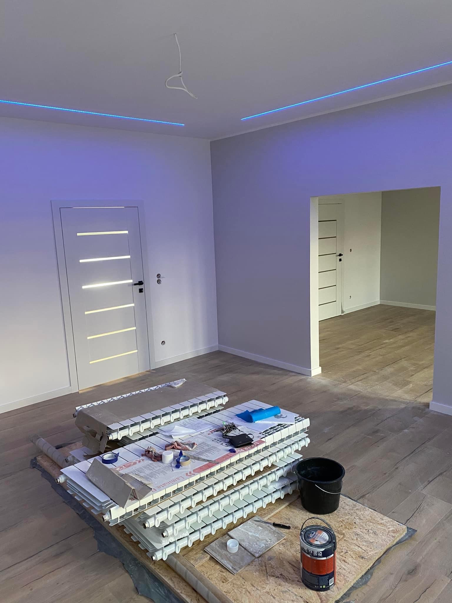 Pokój po remoncie, z nowymi drzwiami z podświetlanymi paskami, świeżo pomalowanymi ścianami, listwami LED pod sufitem i przygotowanymi grzejnikami do montażu na podłodze.