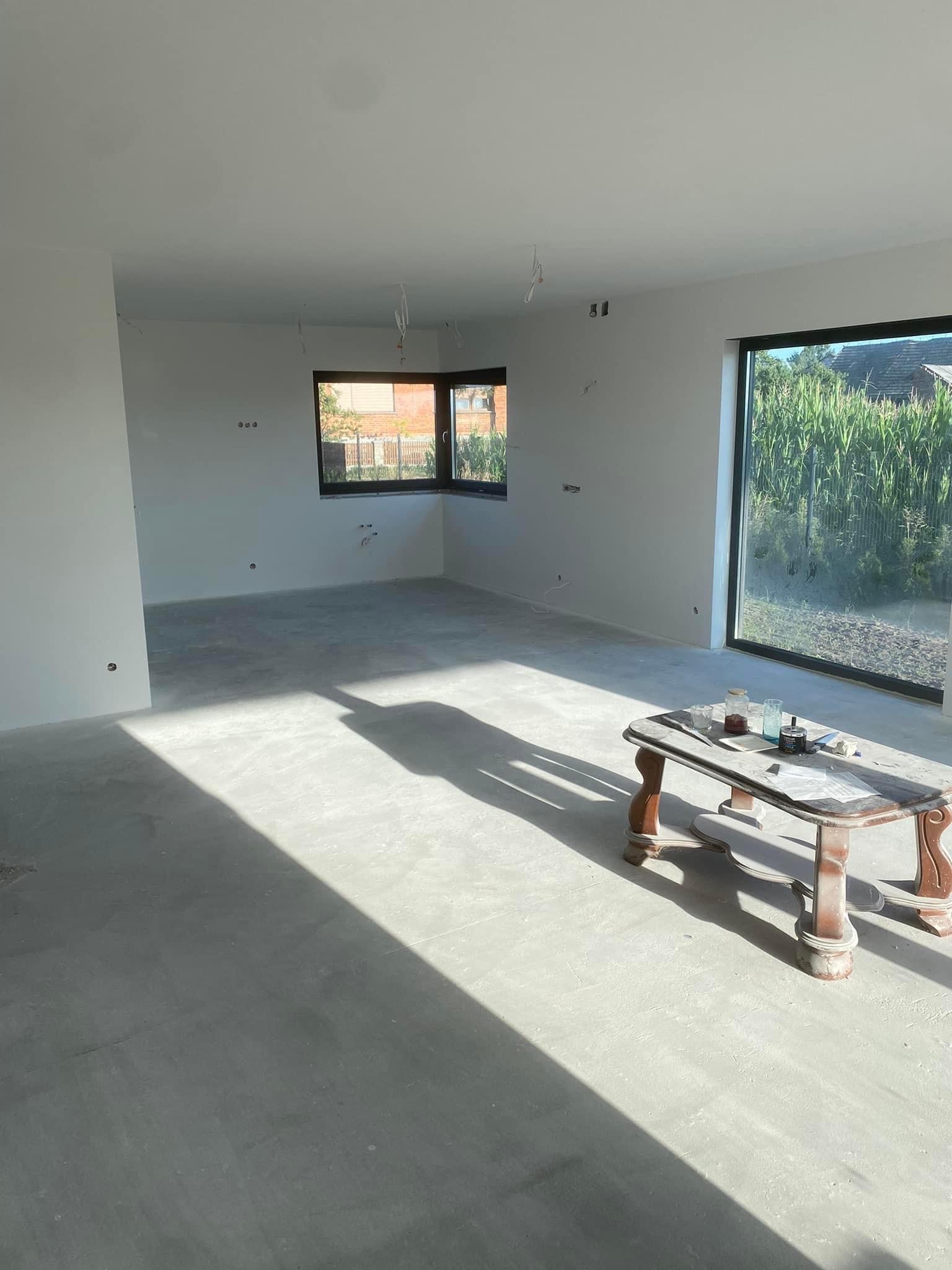 Surowe wnętrze po szpachlowaniu, z widocznymi przewodami elektrycznymi, oknami z widokiem na zieleń i stolikiem z narzędziami malarskimi oświetlonym mocnym światłem słonecznym.
