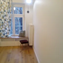 Jasne wnętrze pokoju po remoncie: parkiet, białe ściany, okno z zasłoną w geometryczne wzory, siedzisko pod oknem, krzesło z bordowym siedziskiem i grzejnik.