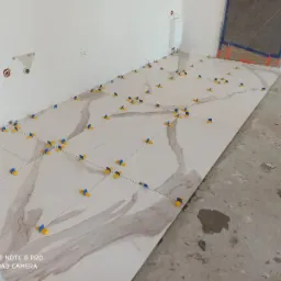 Układanie płytek podłogowych z wzorem marmuru, z użyciem żółto-niebieskich krzyżyków dystansowych, widoczne przygotowane podłoże i fragment ściany z instalacją elektryczną.