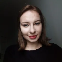 Portret młodej kobiety z kr&oacute;tkimi brązowymi włosami, uśmiechniętej, w czarnym swetrze na tle ściany w odcieniach szarości.