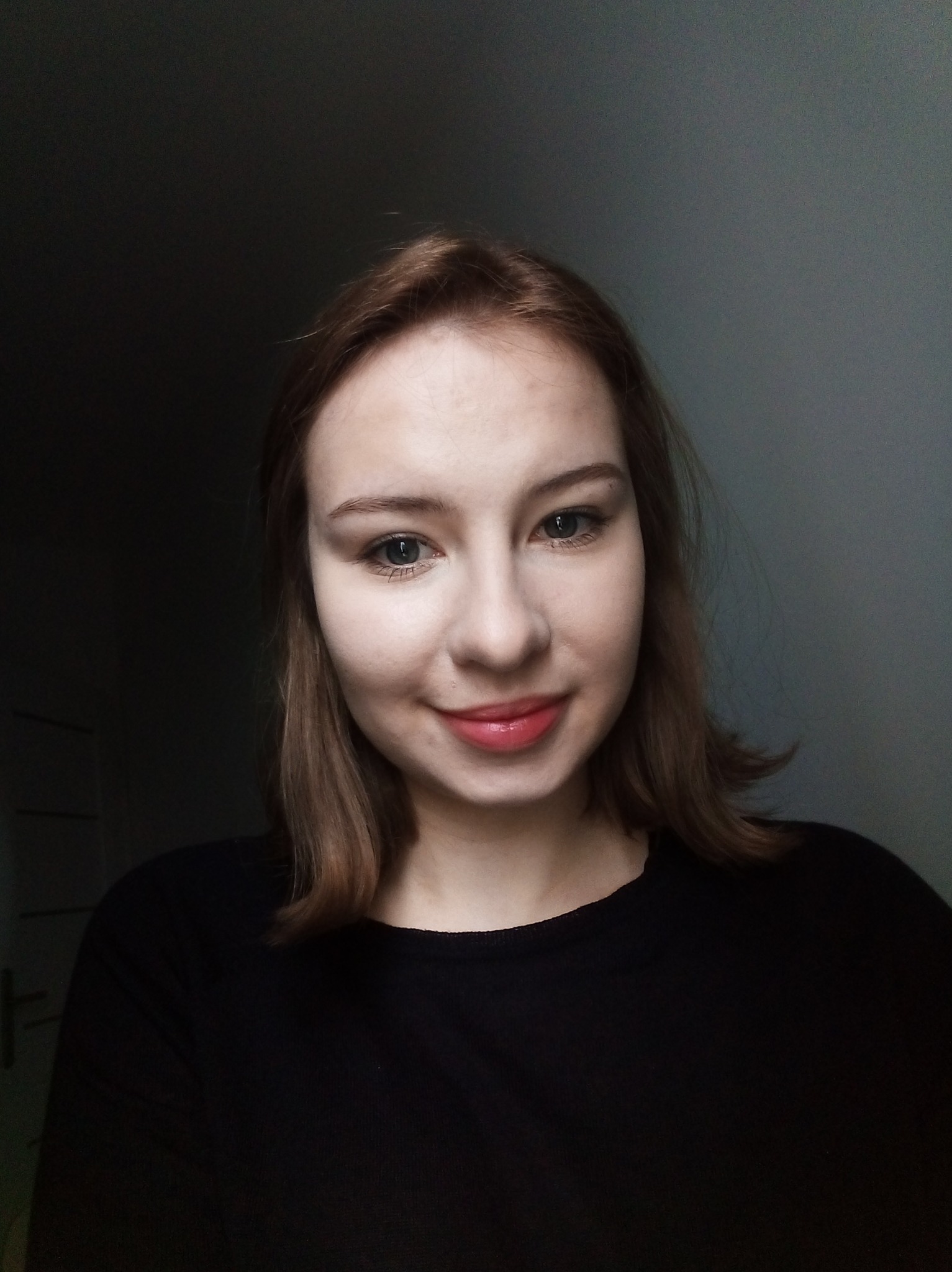 Portret młodej kobiety z krótkimi brązowymi włosami, uśmiechniętej, w czarnym swetrze na tle ściany w odcieniach szarości.