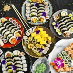 Różnorodny zestaw sushi i rolek na kilku talerzach, udekorowany jadalnymi kwiatami, podany na ciemnym tle z pałeczkami i wasabi.