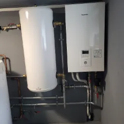 Biały zasobnik ciepłej wody Galmet i wiszący moduł Panasonic Aquarea Smart Cloud z licznymi rurami i zaworami, zamontowane na szarej ścianie w pomieszczeniu technicznym.