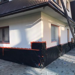 Alp Pro Prace Wysokościowe - Przygotowania do prac elewacyjnych: ściana budynku zabezpieczona folią i taśmą malarską, okna zakryte folią, drabina oparta o ścianę, widoczny fragment dachu z brązowej blachy falistej.