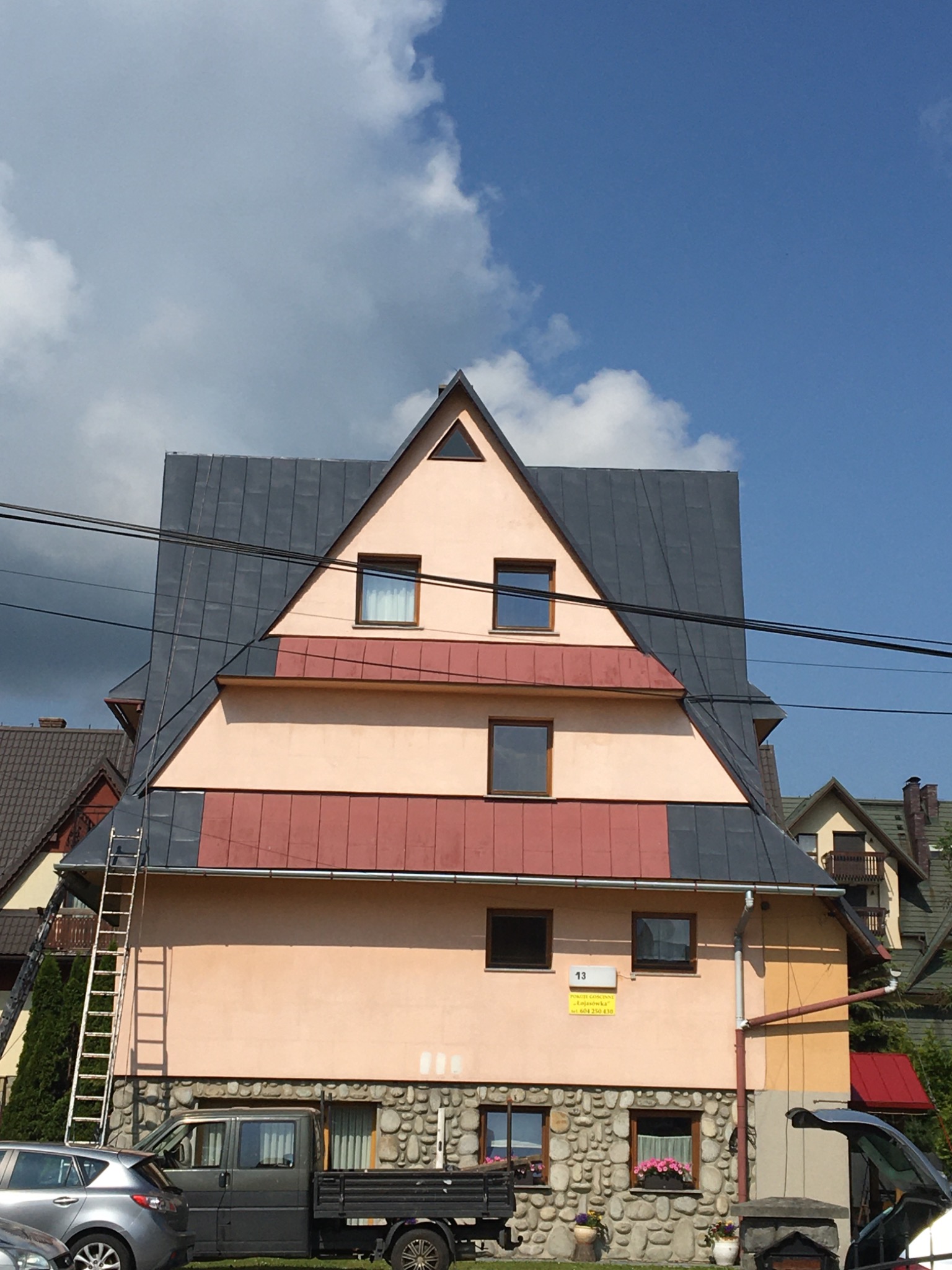 Fasada budynku z nietypowym dachem w kształcie trójkąta, częściowo pokrytym blachą, z drabiną opartą o ścianę i zaparkowanymi samochodami.