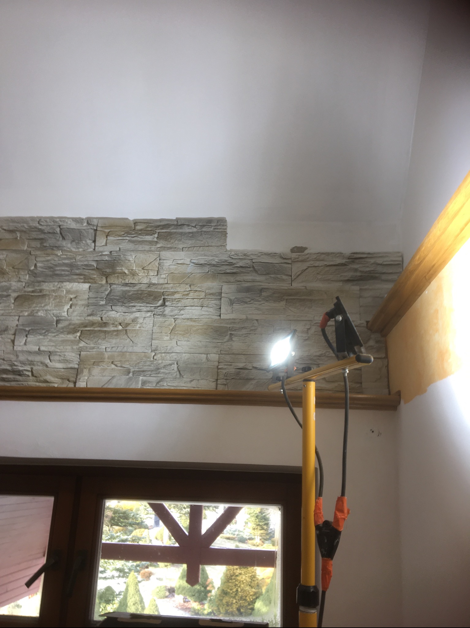 Montaż dekoracyjnej ściany z imitacji kamienia wewnątrz pomieszczenia, oświetlonej lampą roboczą na statywie, widok z dołu na fragment okna.