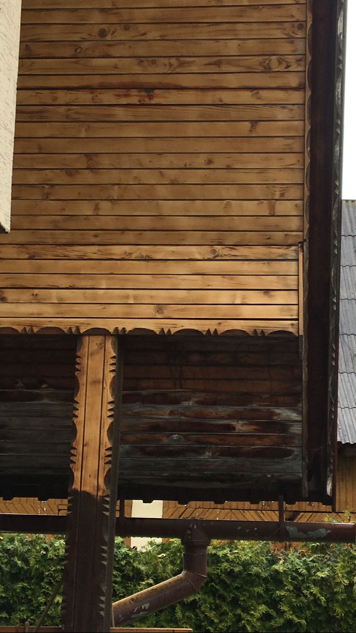 Fragment drewnianej elewacji domu z dekoracyjnymi elementami, widoczny spust rynnowy i fragment dachu sąsiedniego budynku.