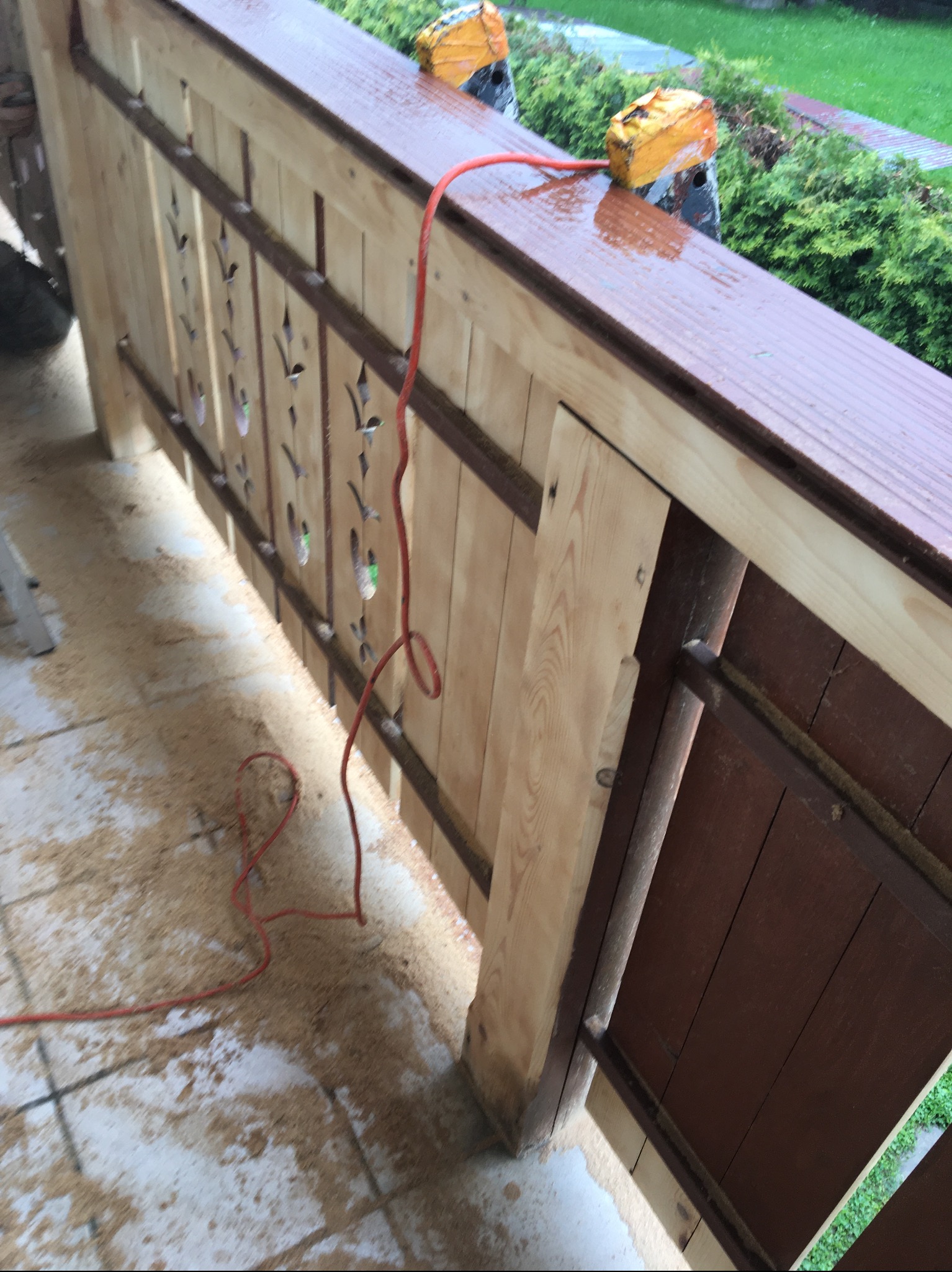 Drewniana balustrada balkonu w trakcie renowacji, z widocznymi śladami po obróbce drewna, zabezpieczona folią malarską, z częściowo odnowionymi elementami i czerwonym kablem zasilającym...