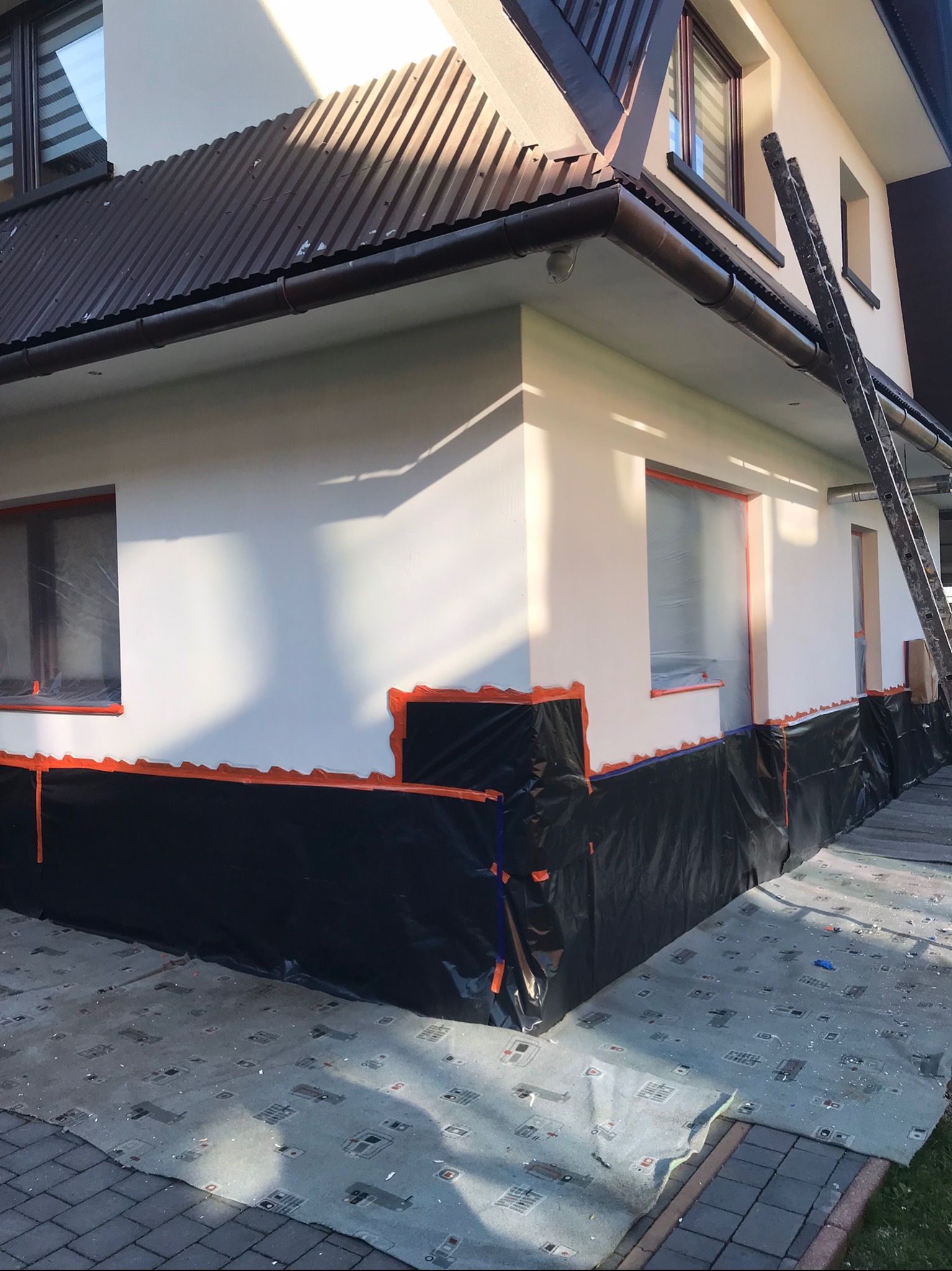 Przygotowania do prac elewacyjnych: ściana budynku zabezpieczona folią i taśmą malarską, okna zakryte folią, drabina oparta o ścianę, widoczny fragment dachu z brązowej blachy falistej.